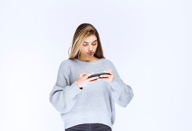 Fille en sweat-shirt gris tenant un smartphone noir et écrivant un message ou un commentaire.