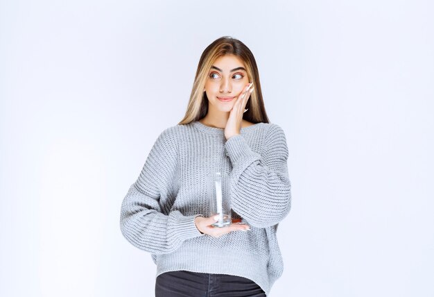 Fille en sweat-shirt gris tenant un verre d'eau pure et faisant le visage doutant.