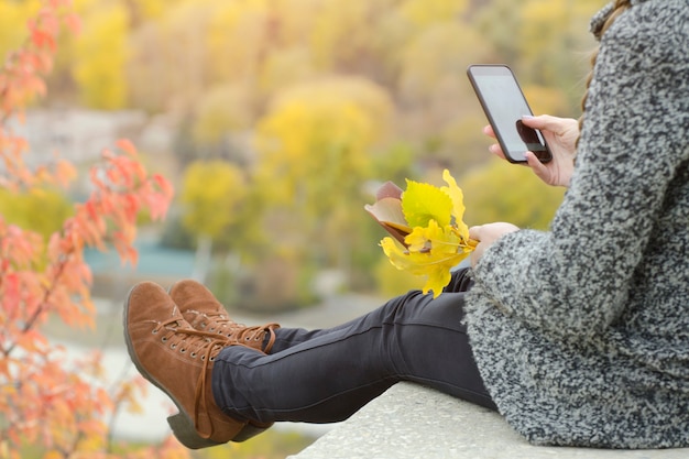 Une fille avec un téléphone est assise sur une colline, des feuilles jaunes à la main. l'automne en arrière-plan
