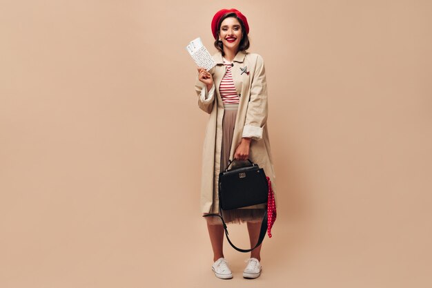 Fille en tenue élégante pose avec sac à main et billets. Belle jeune femme en jupe longue beige et sourires de manteau sur fond isolé.