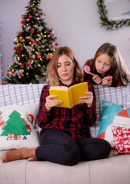 Fille tient la canne en bonbon et regarde sa mère lire un livre assis sur un canapé et profiter du temps de Noël à la maison
