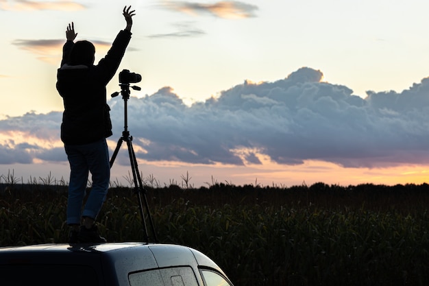 La fille sur le toit de la voiture photographie le coucher de soleil avec un trépied