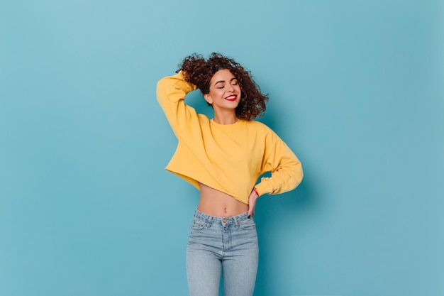 Une fille touche ses cheveux bouclés et sourit les yeux fermés Photo d'une femme avec du rouge à lèvres vêtue d'un pull jaune et d'un pantalon en jean sur fond bleu