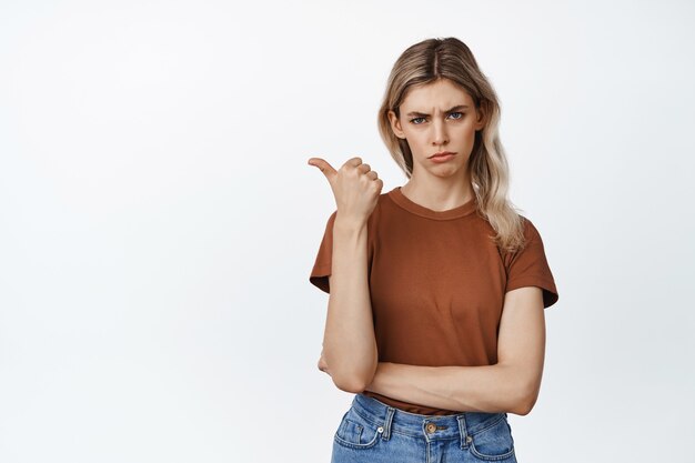 Fille triste et de mauvaise humeur pointant vers la gauche, regardant avec une expression de visage sceptique, exprime sa déception sur blanc