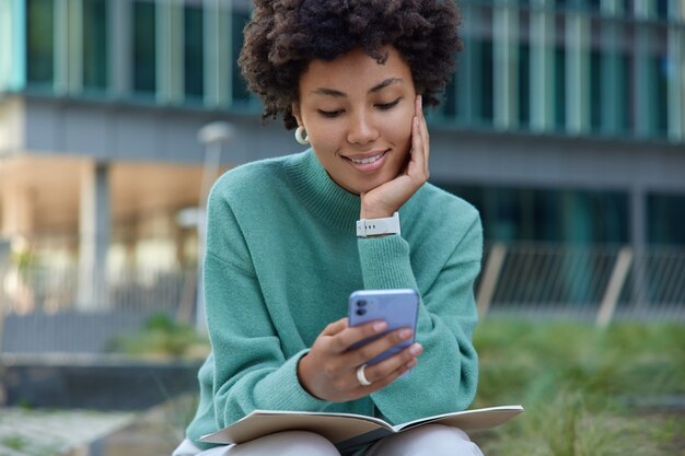 une fille utilise un téléphone portable fait une pause après avoir appris le matériel et se prépare aux poses d'examen avec un journal à l'extérieur dans un cadre urbain porte un pull décontracté