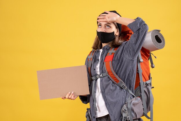 fille de voyageur avec masque noir et sac à dos tenant un carton mettant la main sur sa tête