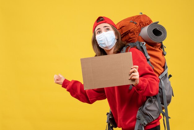 Fille de voyageur vue de face avec sac à dos et masque tenant le carton pointant vers l'arrière