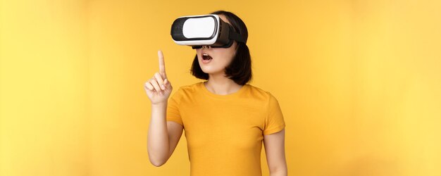 Fille en VR Belle jeune femme asiatique utilisant des lunettes de réalité virtuelle et jouant à discuter virtuellement debout sur fond jaune