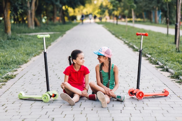 Filles assis sur le trottoir avec leurs scooters poussés sur le trottoir dans le parc