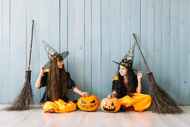 Photo gratuite filles en costumes d'halloween avec des chapeaux pointus et des balais assis près d'une clôture