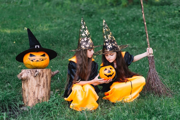 Photo gratuite filles en costumes de sorcière assis sur l'herbe, regardant la citrouille