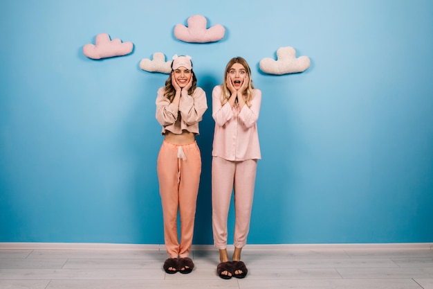 Filles émotionnelles posant en pyjama rose Prise de vue en studio de deux amis qui rient debout sur fond bleu avec de faux nuages