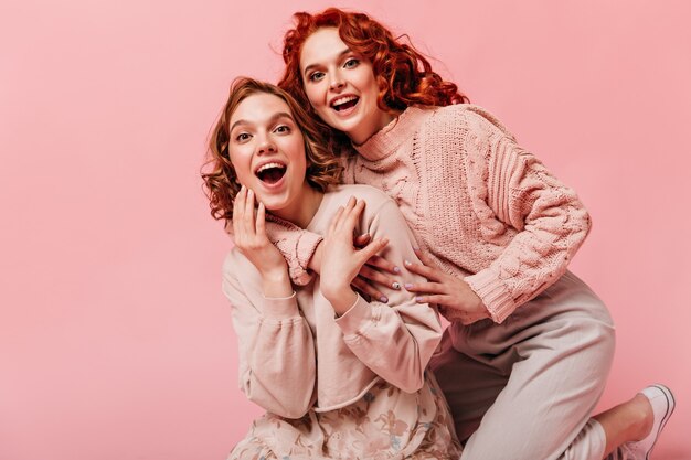 Filles excitées embrassant avec un sourire sincère. Vue de face d'amis heureux riant sur fond rose.