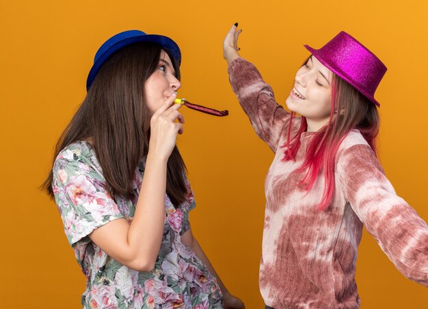 Des filles heureuses portant un chapeau de fête soufflant un sifflet de fête se regardent isolées sur un mur orange