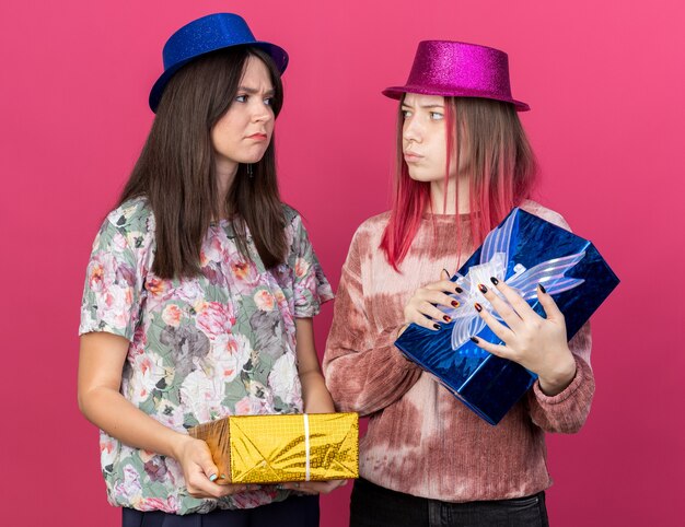 Des filles mécontentes portant un chapeau de fête tenant des coffrets cadeaux se regardent isolées sur un mur rose