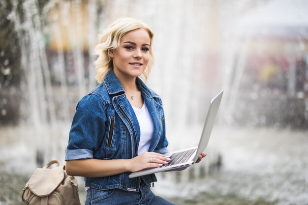 Fit fille blonde étudiante travaille sur son ordinateur portable près de la fontaine dans la ville dans la journée