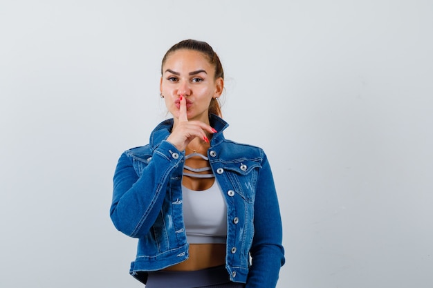 Fit woman in crop top, veste en jean, leggings montrant un geste de silence et l'air sérieux, vue de face.