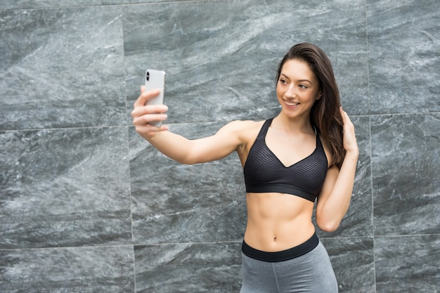 Fitness beauté femme devant le mur extérieur prendre selfie avec smartphone dans la ville après l'entraînement à partager dans les réseaux sociaux