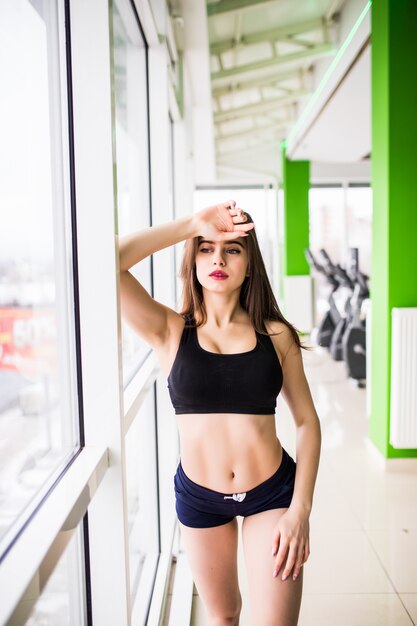 Fitness jeune beauté aux grands yeux verts corps en forme solide longs cheveux bruns pose dans la salle de gym devant les fenêtres