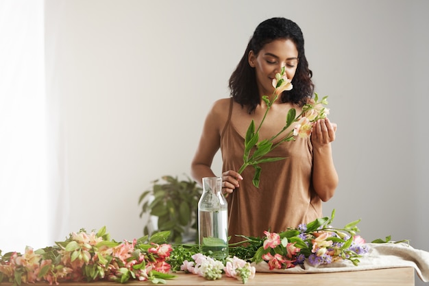 Fleuriste de belle femme africaine aux yeux fermés souriant reniflant des fleurs d'alstroemeria sur mur blanc.