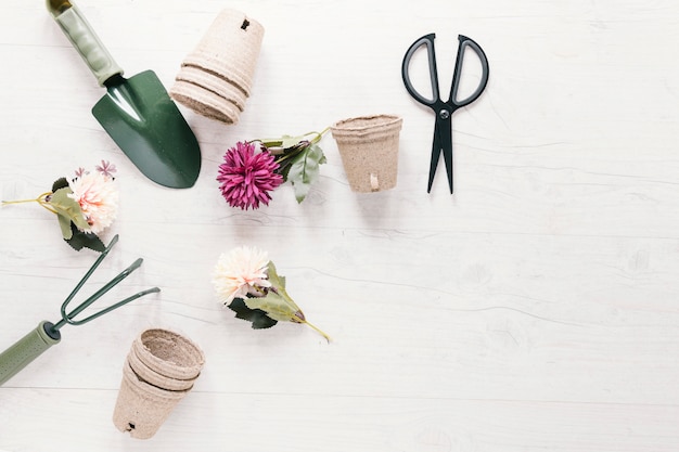 Fleurs artificielles; pot de tourbe et outils de jardinage disposés en forme circulaire avec des ciseaux sur une table blanche