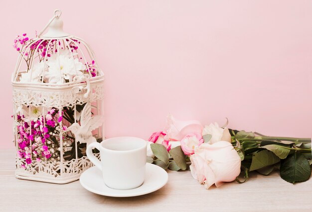 Fleurs en cage antique; tasse; soucoupe et roses sur une table en bois sur fond rose