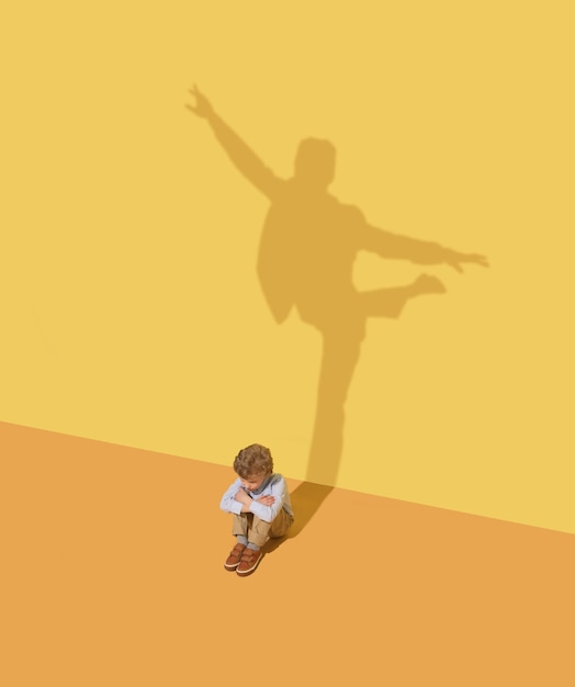 Photo gratuite la flexibilité. concept d'enfance et de rêve. image conceptuelle avec enfant et ombre sur le mur jaune du studio. petit garçon veut devenir danseur de ballet, artiste de théâtre ou homme d'affaires, homme de bureau.