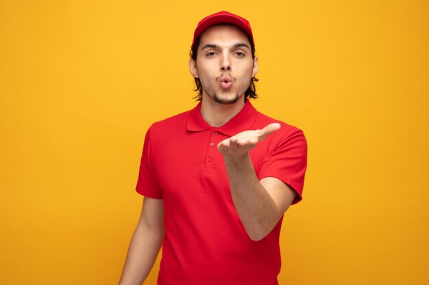 flirty jeune livreur portant l'uniforme et la casquette regardant la caméra soufflant un baiser isolé sur fond jaune