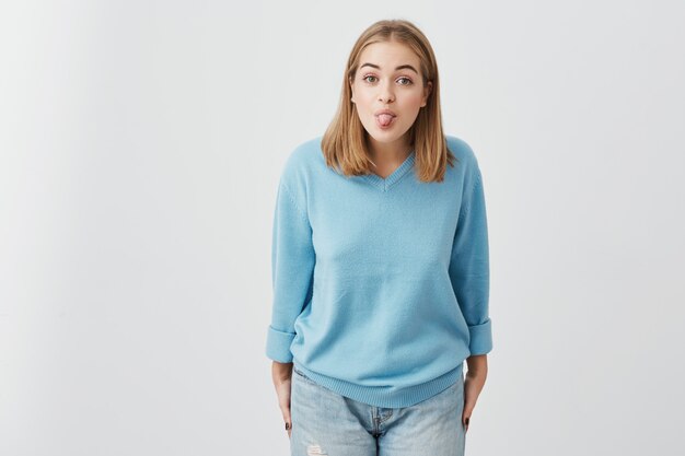 Fond de jolie fille drôle qui sort sa langue. Tête de charmante jeune femme blonde aux cheveux blonds tout en s'amusant à l'intérieur. Gens, style de vie, jeunesse et bonheur