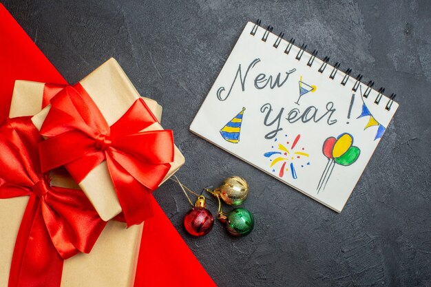 Fond de Noël avec de beaux cadeaux avec ruban en forme d'arc sur une serviette rouge et un cahier avec des dessins de nouvel an sur une table sombre