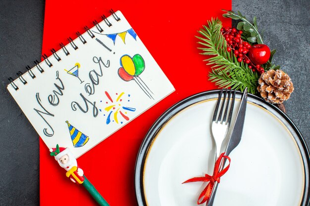Fond de Noël avec des couverts avec ruban rouge sur une assiette à dîner accessoires de décoration branches de sapin à côté de l'ordinateur portable avec un stylo sur une serviette rouge