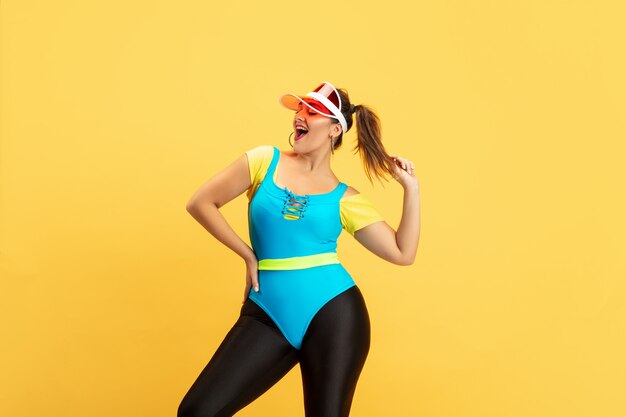 Formation du jeune modèle féminin de taille plus caucasien sur fond jaune. Espace de copie. Concept de sport, mode de vie sain, corps positif, mode, style. Femme élégante posant confiante en chapeau rouge.