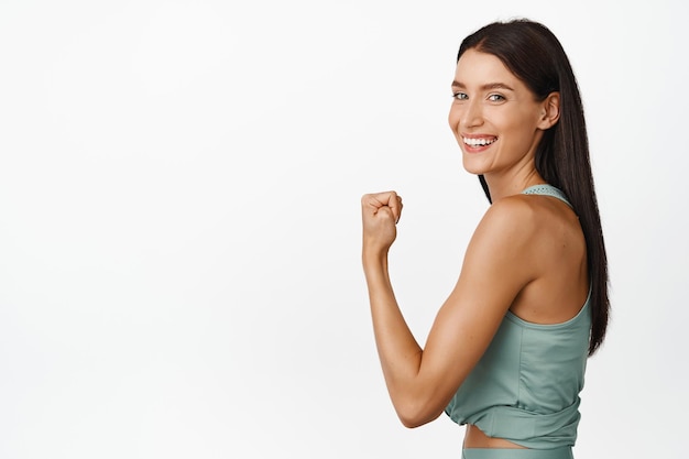 Forte jeune femme de remise en forme montrant les muscles sur le bras fléchissant les biceps et souriant heureux de partager le résultat de l'entraînement debout sur fond blanc