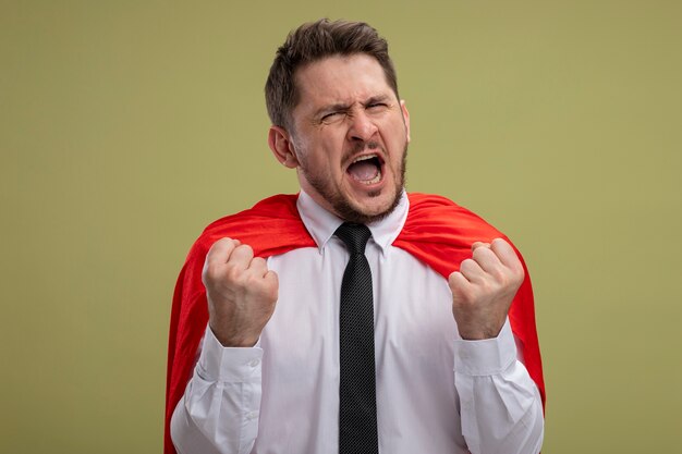 Fou fou et en colère homme d'affaires de super-héros en cape rouge serrant les poings avec une expression agressive va crier sauvage debout sur le mur vert