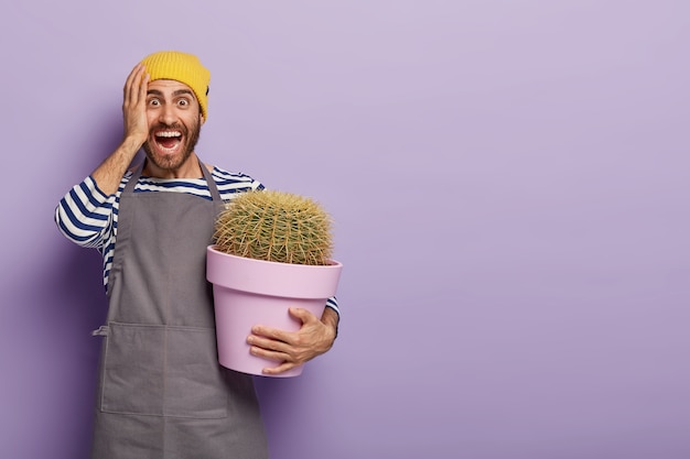 Photo gratuite fou de joie, jeune homme de race blanche détient cactus en pot, vêtu d'un uniforme de travail spécial