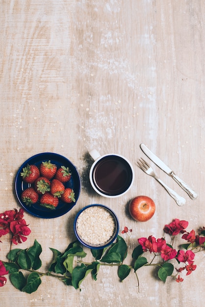 Photo gratuite des fraises; tasse à café; l'avoine; pomme entière et couverts avec des fleurs de bougainvilliers sur fond en bois