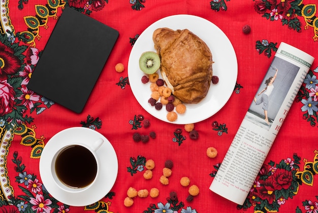 Photo gratuite framboise; kiwi et croissant sur une assiette avec une tasse de café; journal intime et journal enroulé sur nappe