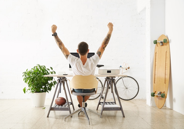 Photo gratuit freelance chanceux tatoué devant son espace de travail, entouré de ses jouets de loisir longboard, vélo vintage et plante verte, étirant sa main dans l'air tout en faisant une pause