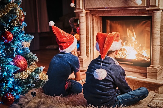 Frère et soeur portant des chapeaux du Père Noël se réchauffant à côté d'une cheminée dans un salon décoré pour Noël.