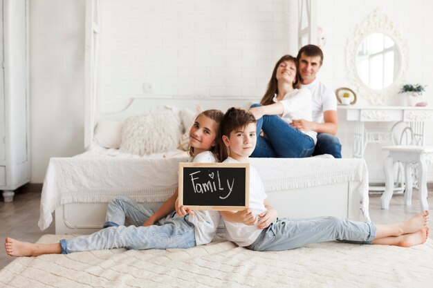 Frère et soeur tenant l'ardoise avec le texte de la famille assis sur un tapis devant leurs parents