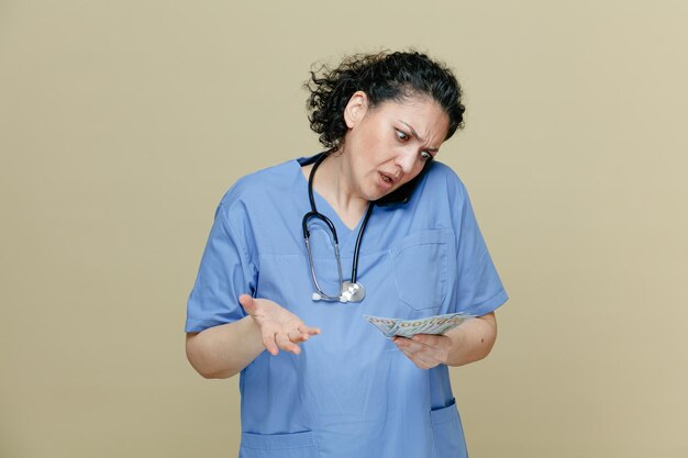 fronçant les sourcils femme médecin d'âge moyen portant l'uniforme et le stéthoscope autour du cou tenant de l'argent regardant vers le bas parler au téléphone montrant la main vide isolée sur fond olive