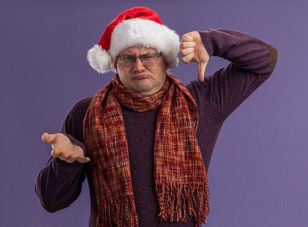 Fronçant les sourcils homme adulte portant des lunettes et bonnet de Noel avec écharpe autour du cou regardant la caméra montrant la main vide et le pouce vers le bas isolé sur fond violet