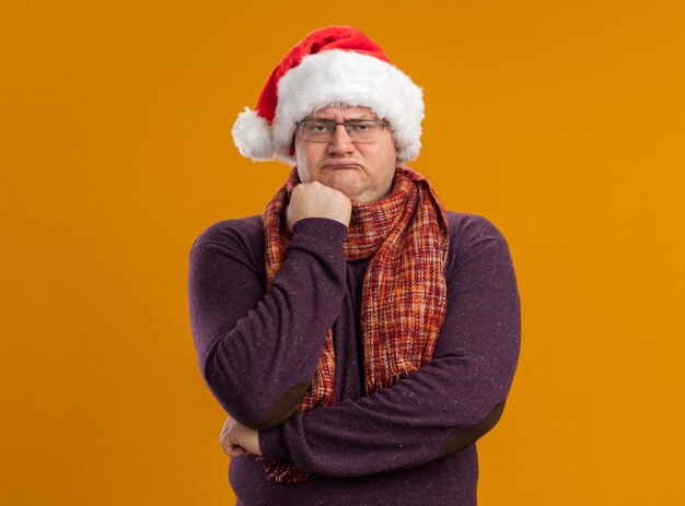 Fronçant les sourcils homme adulte portant des lunettes et bonnet de Noel avec foulard autour du cou en gardant le poing sur le menton regardant la caméra isolée sur fond orange
