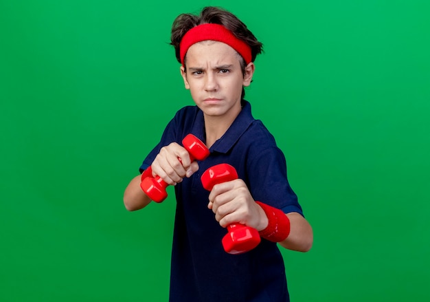 Photo gratuite fronçant les sourcils jeune beau garçon sportif portant un bandeau et des bracelets avec un appareil dentaire tenant des haltères faisant un geste de boxe isolé sur un mur vert avec espace de copie