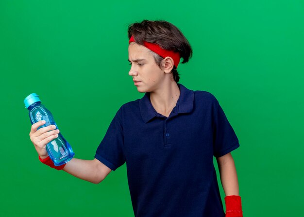 Fronçant les sourcils jeune beau garçon sportif portant un bandeau et des bracelets avec un appareil dentaire tenant et regardant une bouteille d'eau isolée sur un mur vert