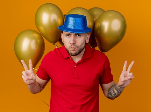 Fronçant les sourcils jeune beau mec de fête portant chapeau de fête debout devant des ballons à l'avant faisant le geste de paix isolé sur un mur orange