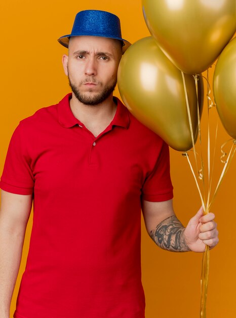 Photo gratuite fronçant les sourcils jeune beau mec de fête portant chapeau de fête tenant des ballons à l'avant isolé sur mur orange