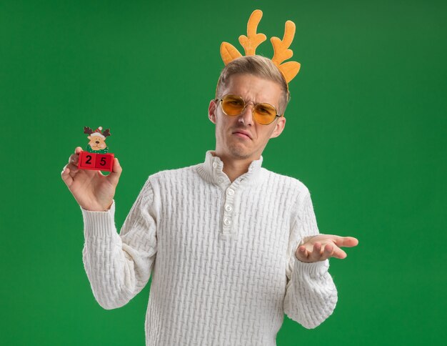 Fronçant les sourcils jeune beau mec portant bandeau de bois de renne avec des lunettes tenant jouet arbre de Noël avec date montrant la main vide isolé sur mur vert
