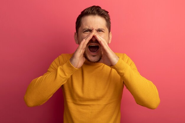 Fronçant les sourcils jeune bel homme blond gardant les mains près de la bouche à la recherche d'appel à haute voix à quelqu'un isolé sur le mur rose