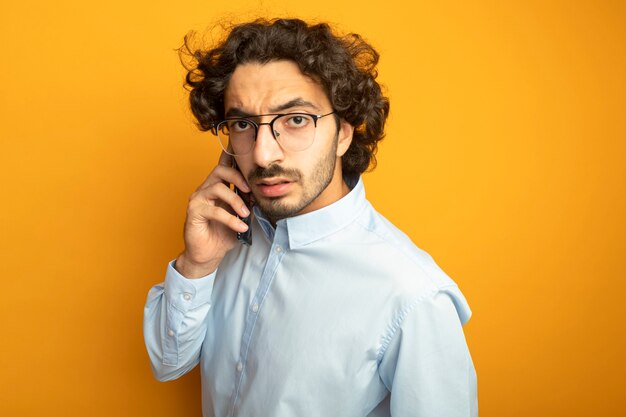 Fronçant les sourcils jeune bel homme caucasien portant des lunettes parler au téléphone en regardant la caméra isolée sur fond orange avec espace de copie